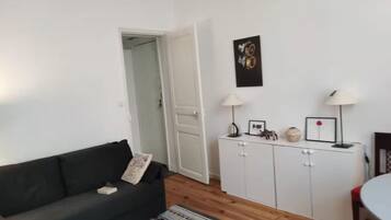 Een woonkamer met een donkergrijze bank, een witte kast met decoratieve objecten, een kamerkleed met dessin, en een deur naar een andere kamer.