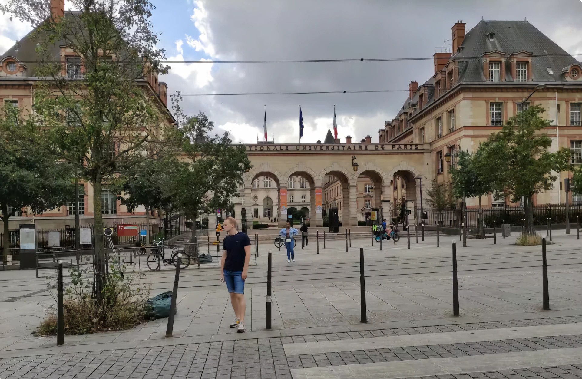 A courtyard with a large building in the background, featuring arched walkways and flags.