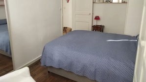 A bedroom with a bed, a door, a small table with a lamp, and a shelf with pictures. - Charming 1‑Bedroom Apartment 500 m from Paris – RER B 2 min – Cité Universitaire (Gentilly)