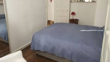 Een slaapkamer met een bed, een deur, een klein tafeltje met een lamp en een plank met ingekaderde foto's.