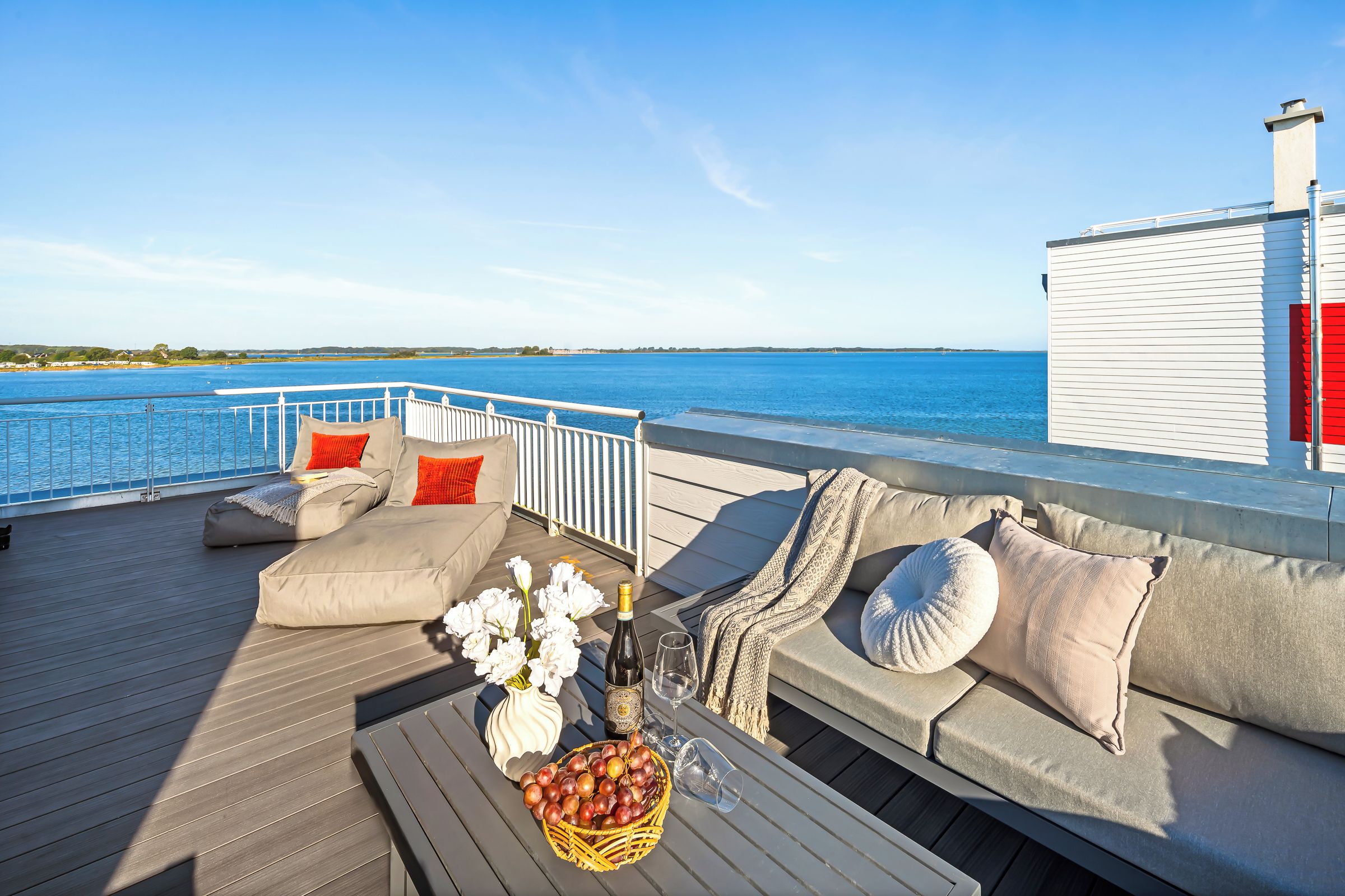 A deck with a sofa, cushions, a table with a bottle and glasses, and a basket of fruit.