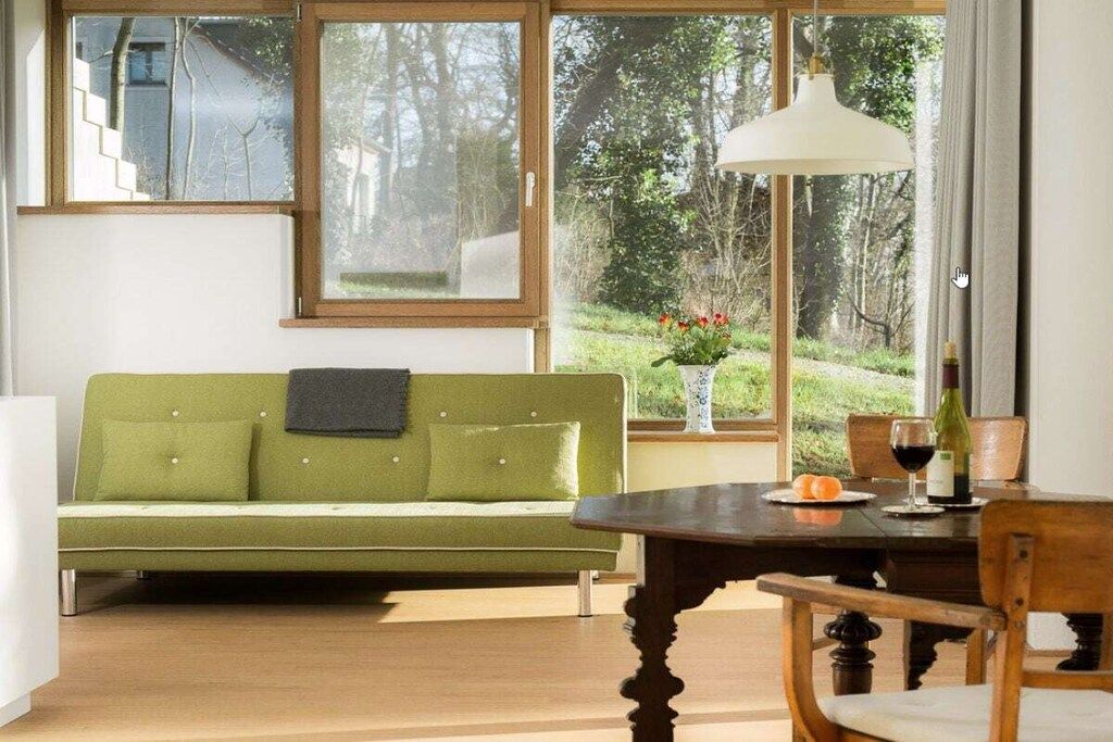 A room with a green sofa, a wooden dining table with chairs, a hanging light fixture, and a window with a view of greenery.
