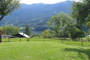 A grassy field with a wooden fence, a building with a sloped roof, and a lake in the background. - Holiday home with 1 bedroom (GRUNDLSEE)