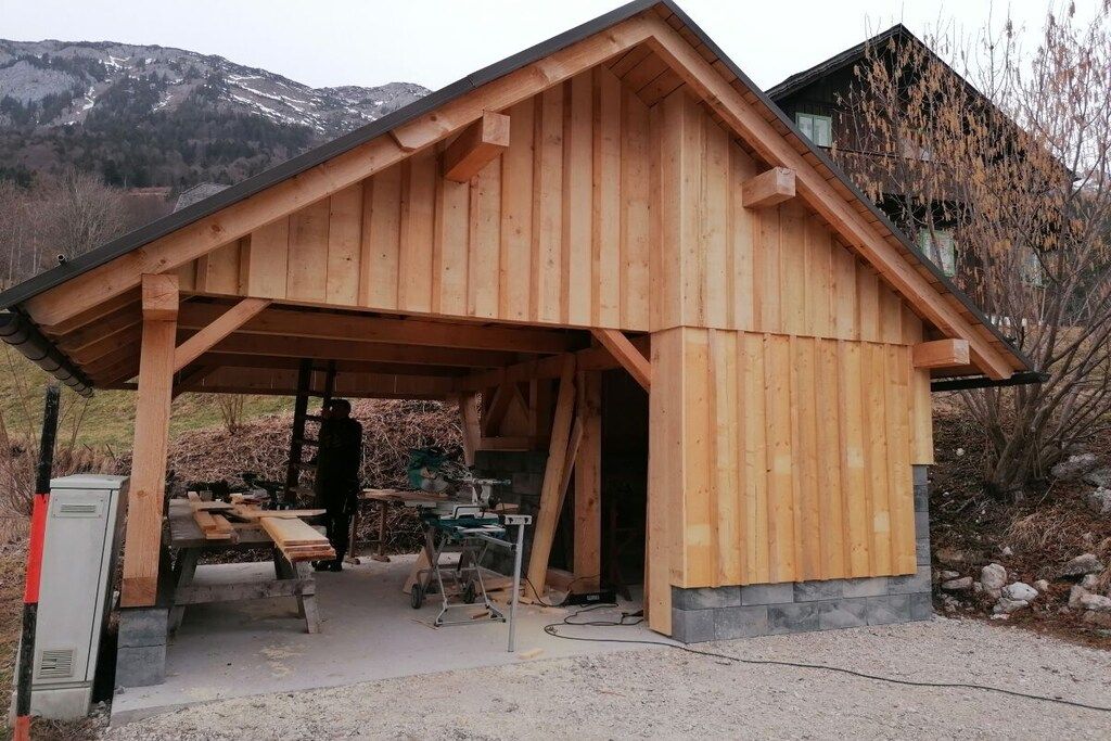 A wooden cabin with a sloped roof, a covered outdoor area with benches, and a mountainous backdrop.