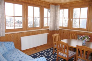 Habitació amb parets de fusta, un radiador blanc, un sofà blau amb dibuixos, una taula de dinar amb cadires i grans finestres amb cortines.