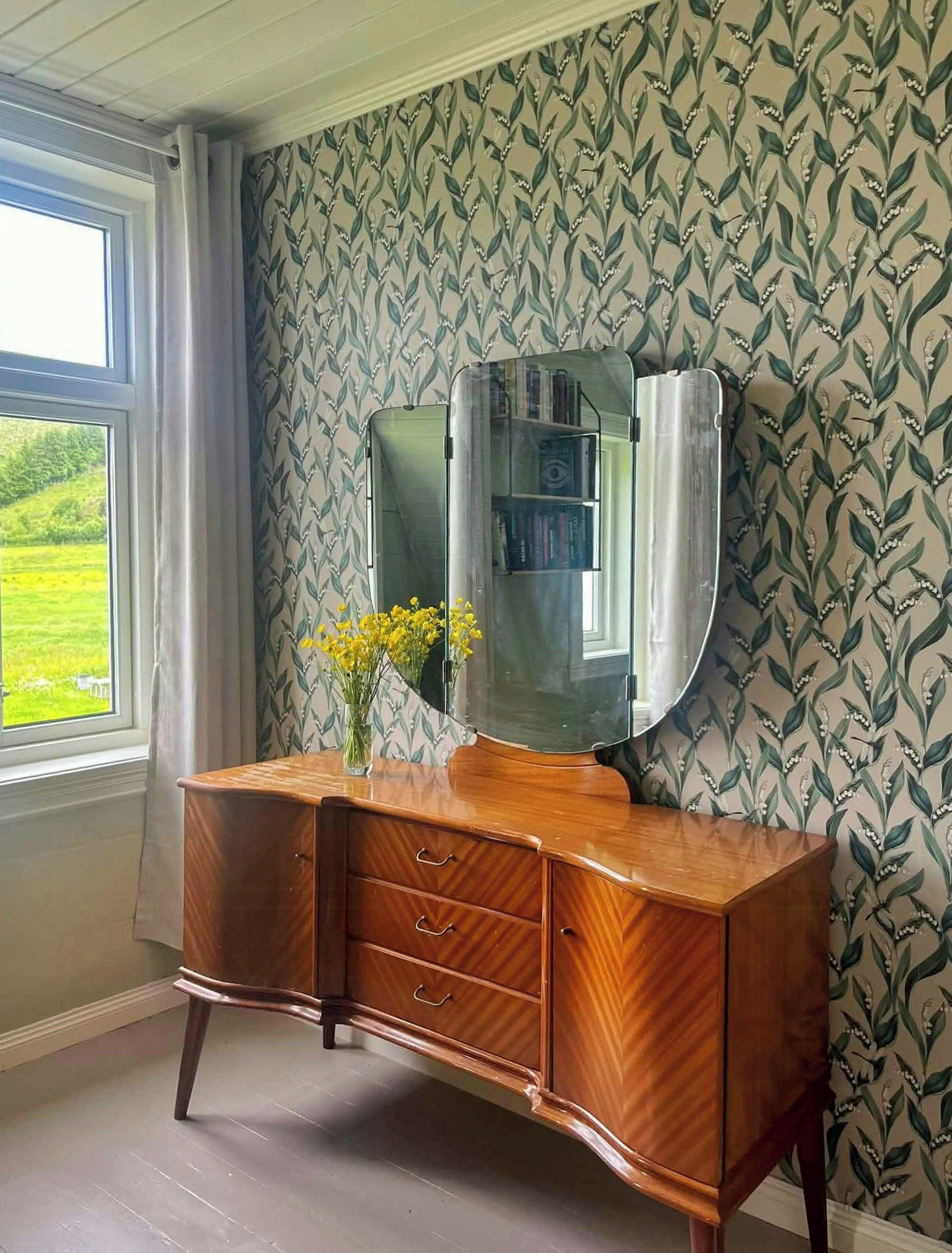 A wooden vanity with a mirror and a vase of yellow flowers against a wallpaper with a leafy pattern.