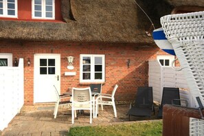 Een stenen huis met rieten dak, een witte deur en een klein terras met tuinmeubilair.