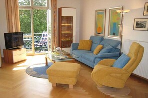 Ein Wohnzimmer mit einer Couch, einem Sessel, einem Hocker und einem Fernseher auf einem Schrank.