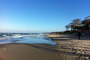 A beach with a person walking on the sand, waves approaching the shore, and a clear sky. - Holiday home in Born with swimming pool (Born)