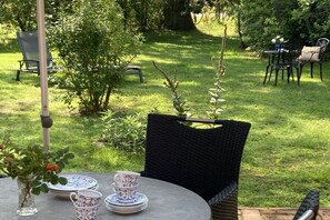 Ein runder Tisch mit Blumen, einem Stapel Tellern und einer Tasse, platziert in einem Garten mit Stühlen und einem Baum im Hintergrund.