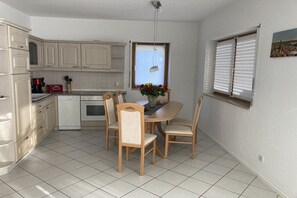 A compact kitchen with white cabinets, a dining table with chairs, a stove, and a window with blinds. - Holiday apartment with 1 bedroom (Bannewitz)