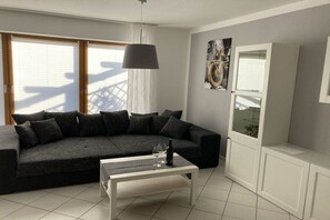 Uma sala de estar moderna com um sofá modular cinza escuro, uma mesa de centro branca e uma janela grande com persianas.