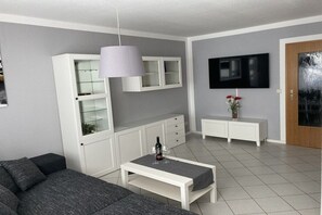 A modern living room with a sofa, a coffee table with a bottle and glasses, a TV mounted on the wall, and a cabinet with glass doors. - Holiday apartment with 1 bedroom (Bannewitz)