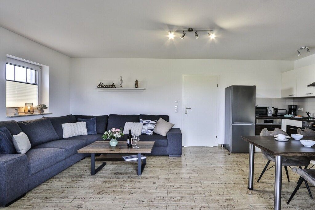 A modern living room with a grey sofa, a wooden coffee table, and a kitchen area with a refrigerator and microwave.