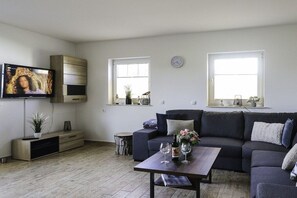 Ein modernes Wohnzimmer mit einem dunkelgrauen Sofa, einem Couchtisch mit einer Vase und Weingläsern, einem flachbildfernseher an der Wand und zwei Fenstern mit Vorhängen.