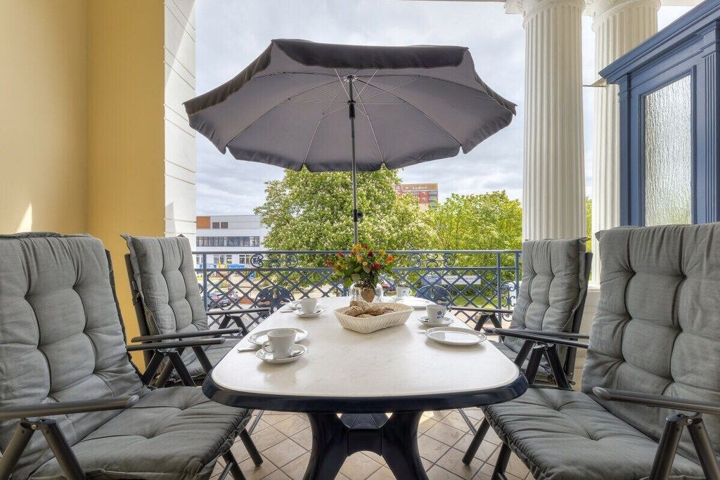Ein Balkon mit rundem Tisch, vier grau gepolsterten Stühlen, einem grauen Sonnenschirm und einer Vase mit Blumen.