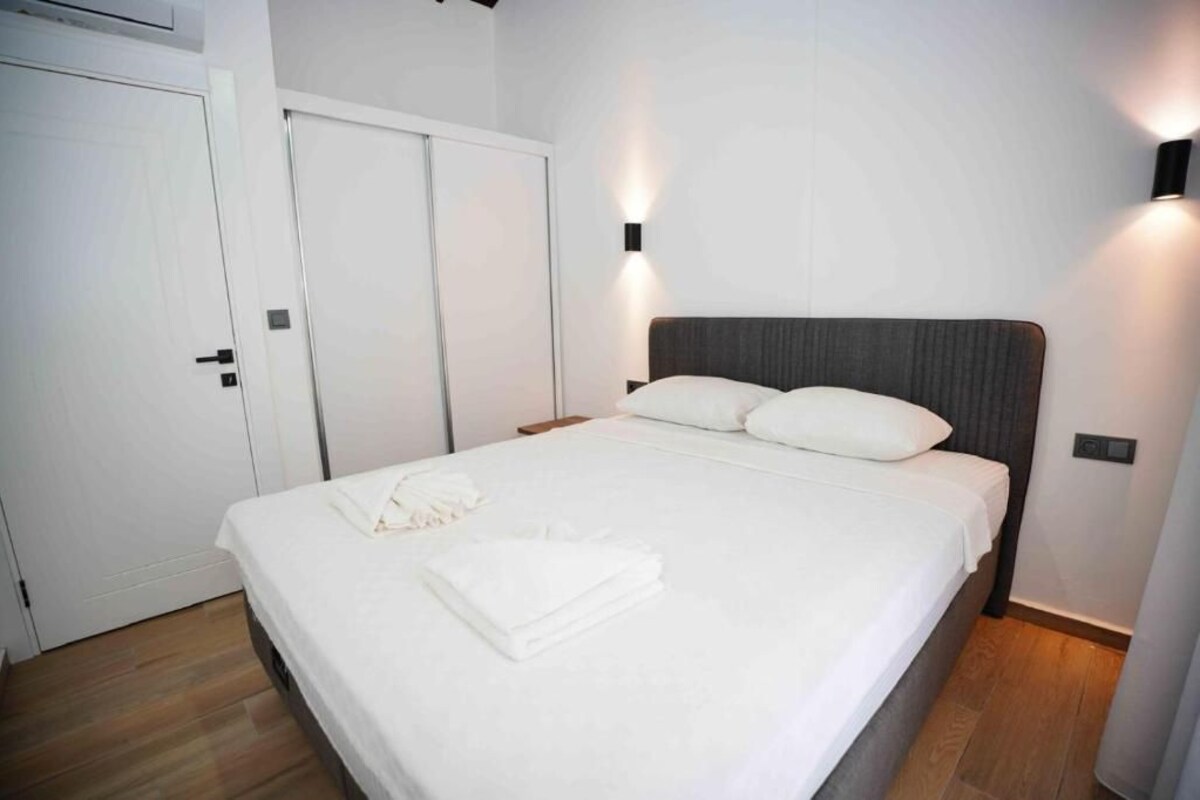 A neatly made bed with white linens and a folded towel, a wooden floor, and a white door.