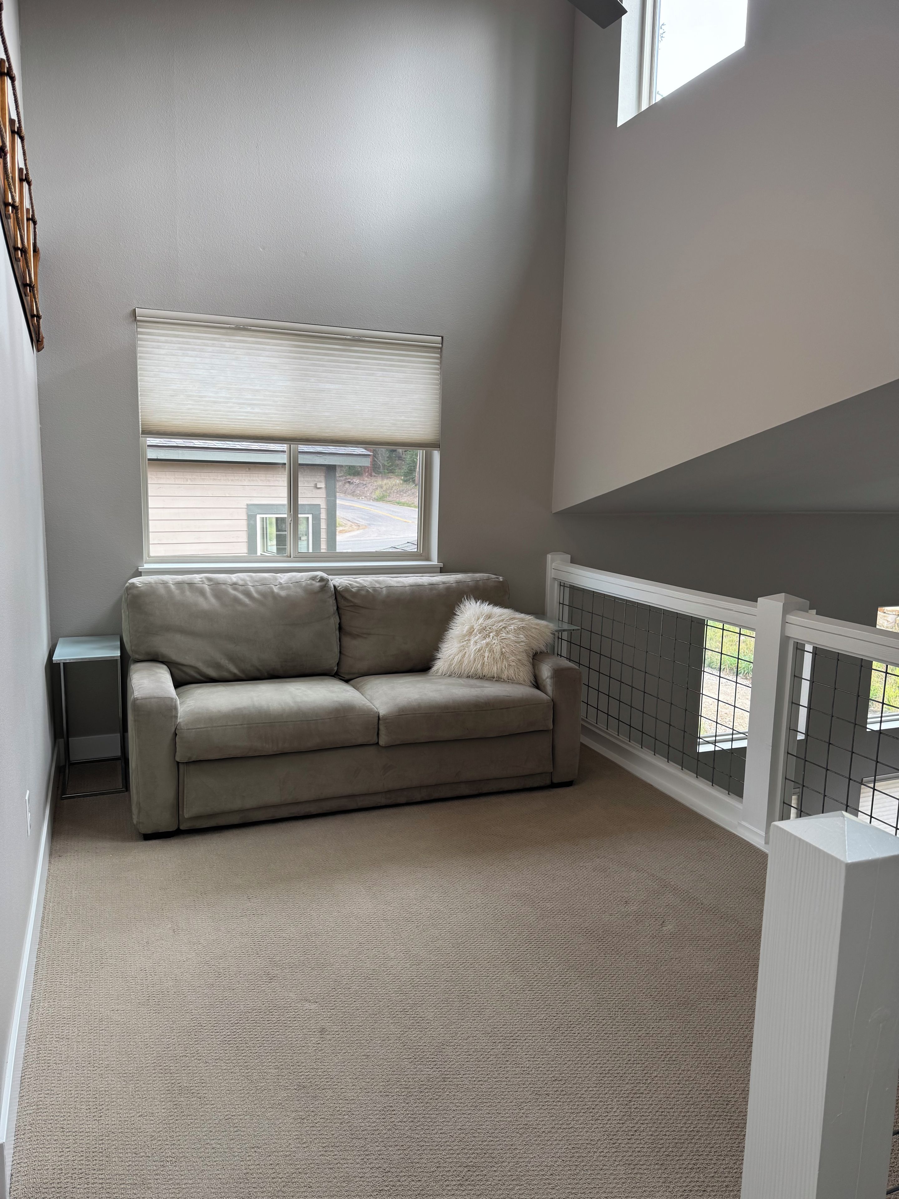 A room with a beige sofa, a small side table, a window with blinds, and a view of the outside.
