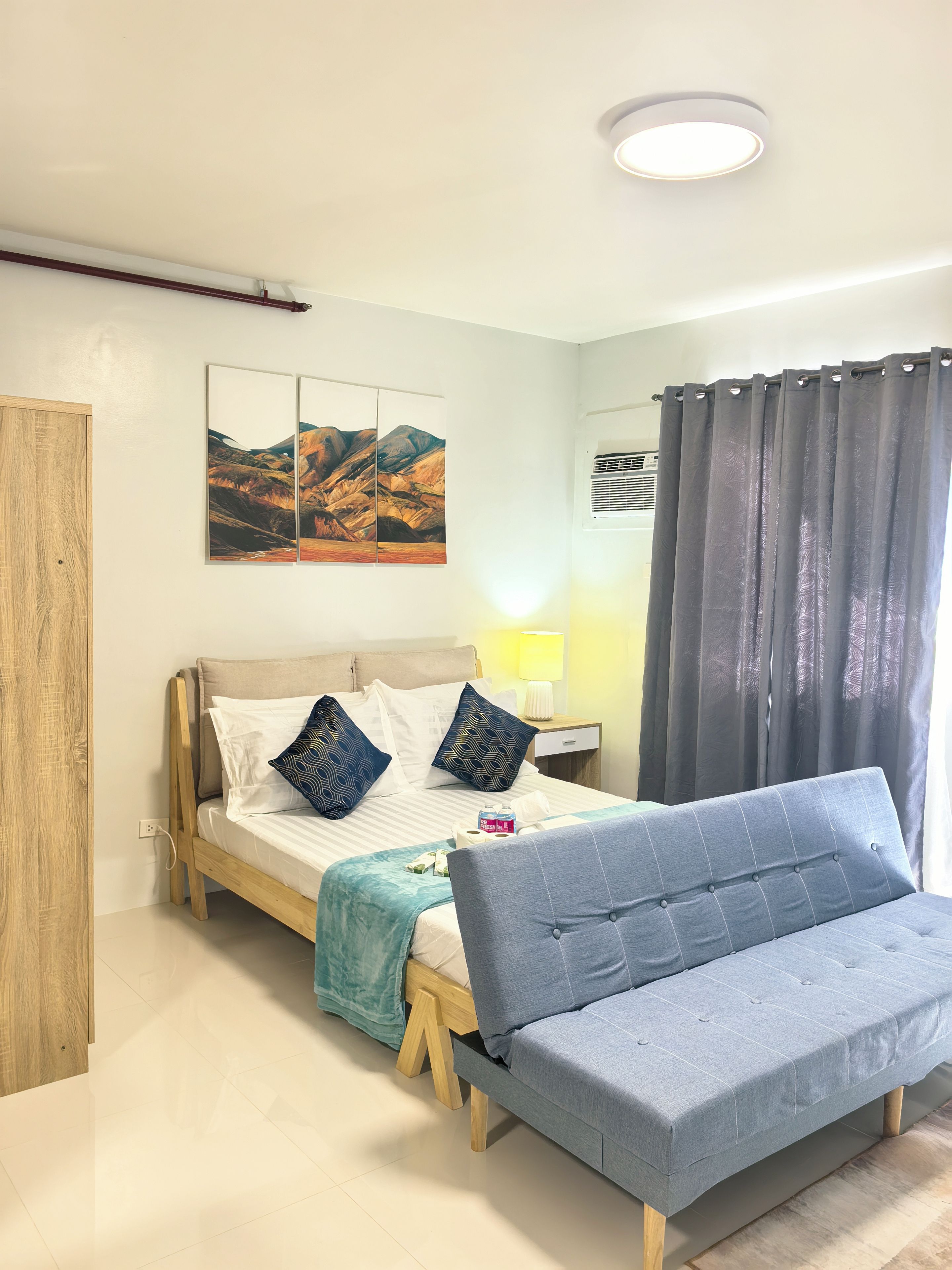 Ein modernes Schlafzimmer mit einem Bett, einem Nachttisch mit Lampe, einer Wand-Klimaanlage und einem großen Fenster mit Vorhängen.