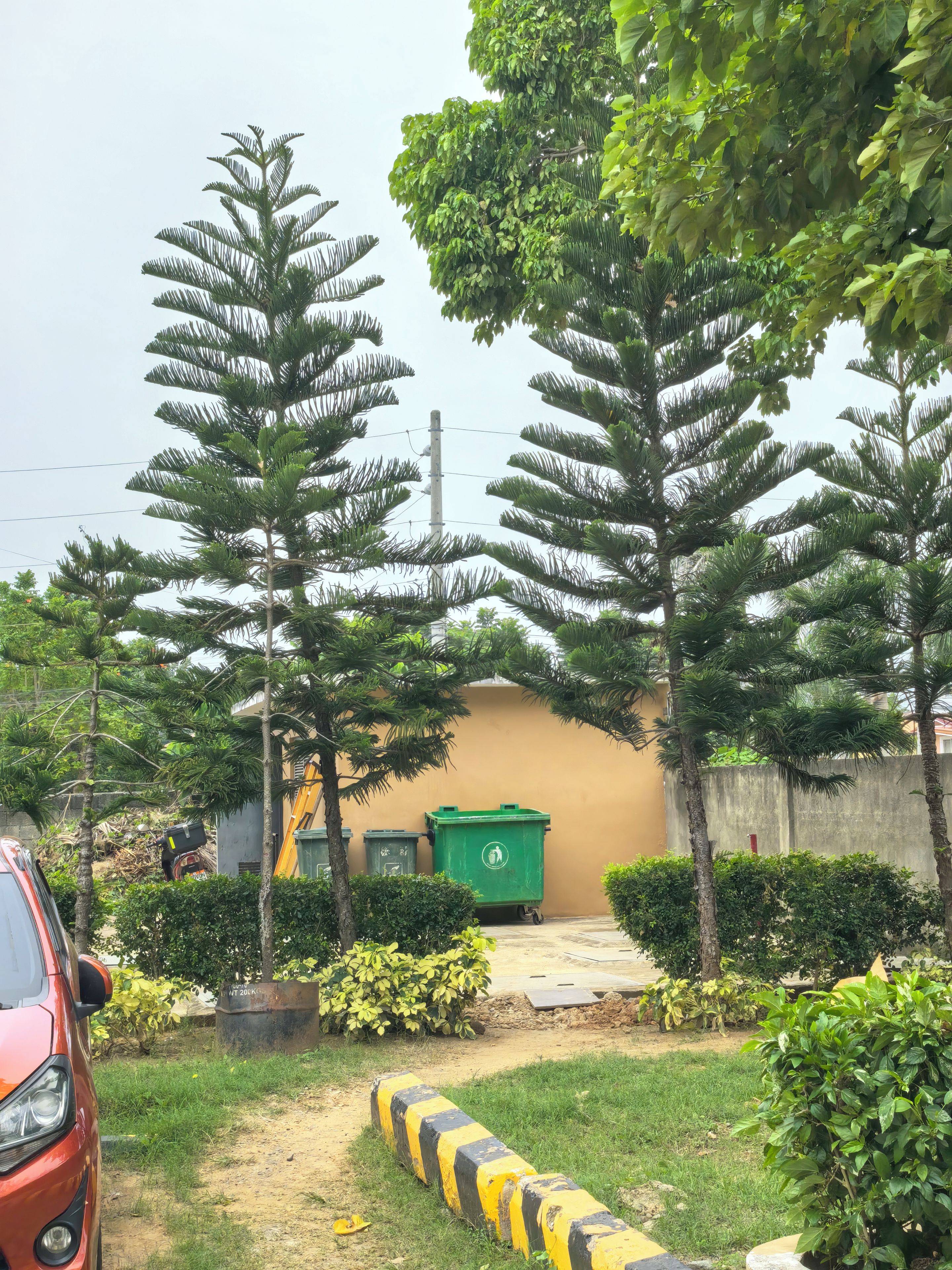 A parking area with a red car, green recycling bins, and a yellow and black striped barrier.