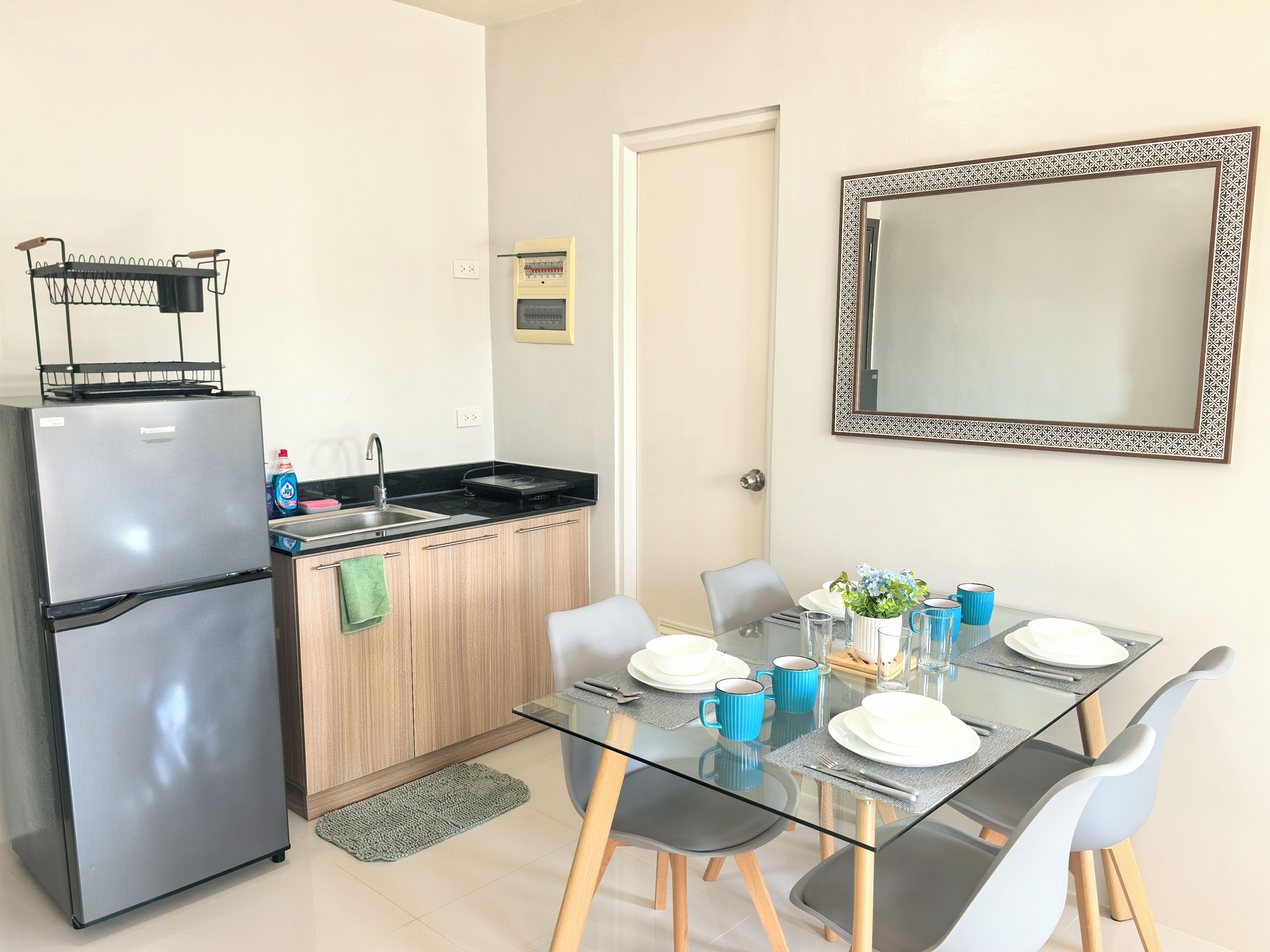 A compact kitchen with a refrigerator, sink, and a small countertop. There is a dining table with chairs and a mirror on the wall.