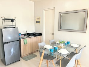 A compact kitchen with a refrigerator, sink, and a small countertop. There is a dining table with chairs and a mirror on the wall. - Camella Verdant Studio,  Gym, Pool Balcony and Near Airport (Puerto Princesa City)