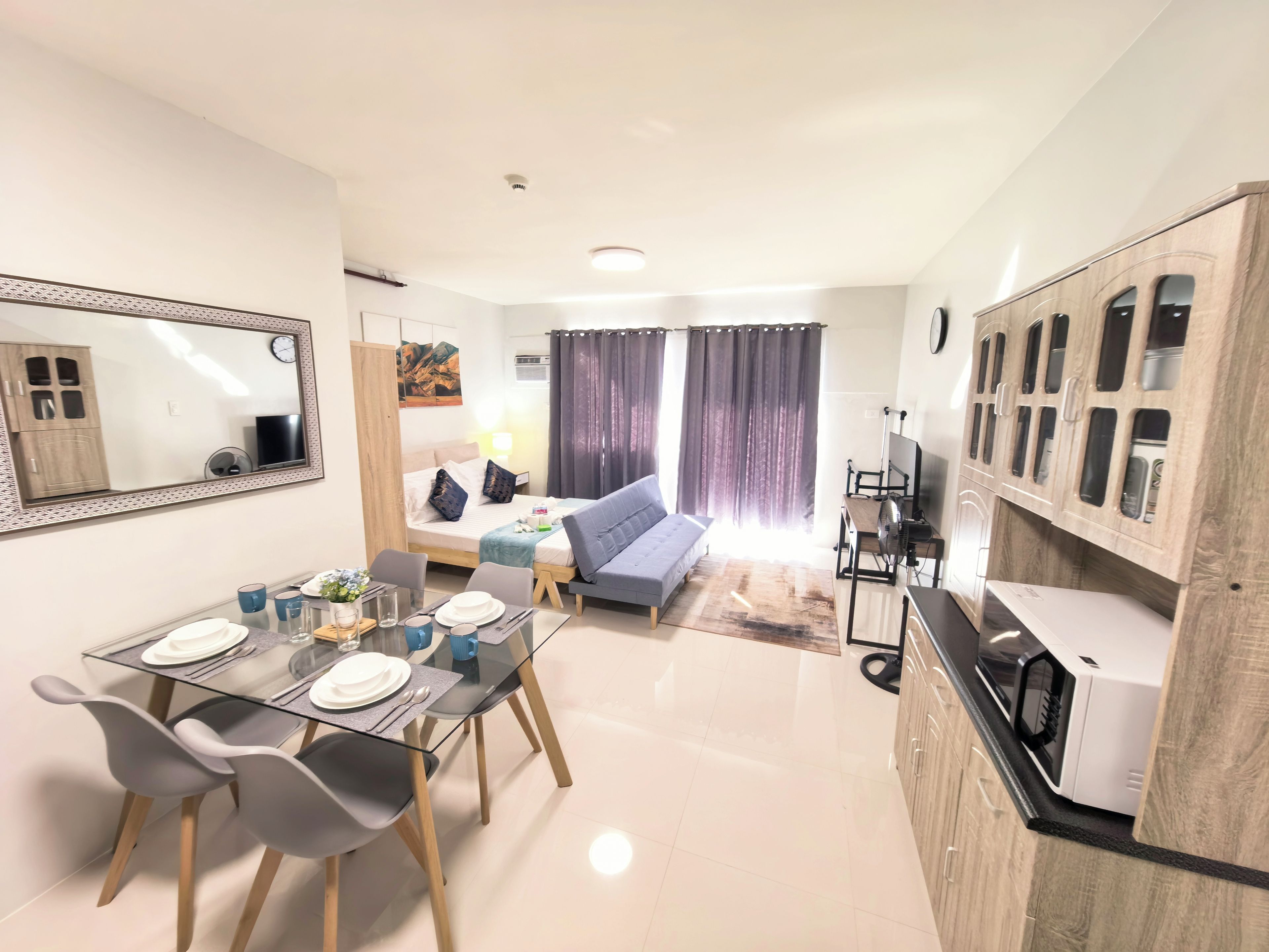 A modern living room with a sofa, a dining table with chairs, a kitchen area with cabinets, and a large mirror on the wall.