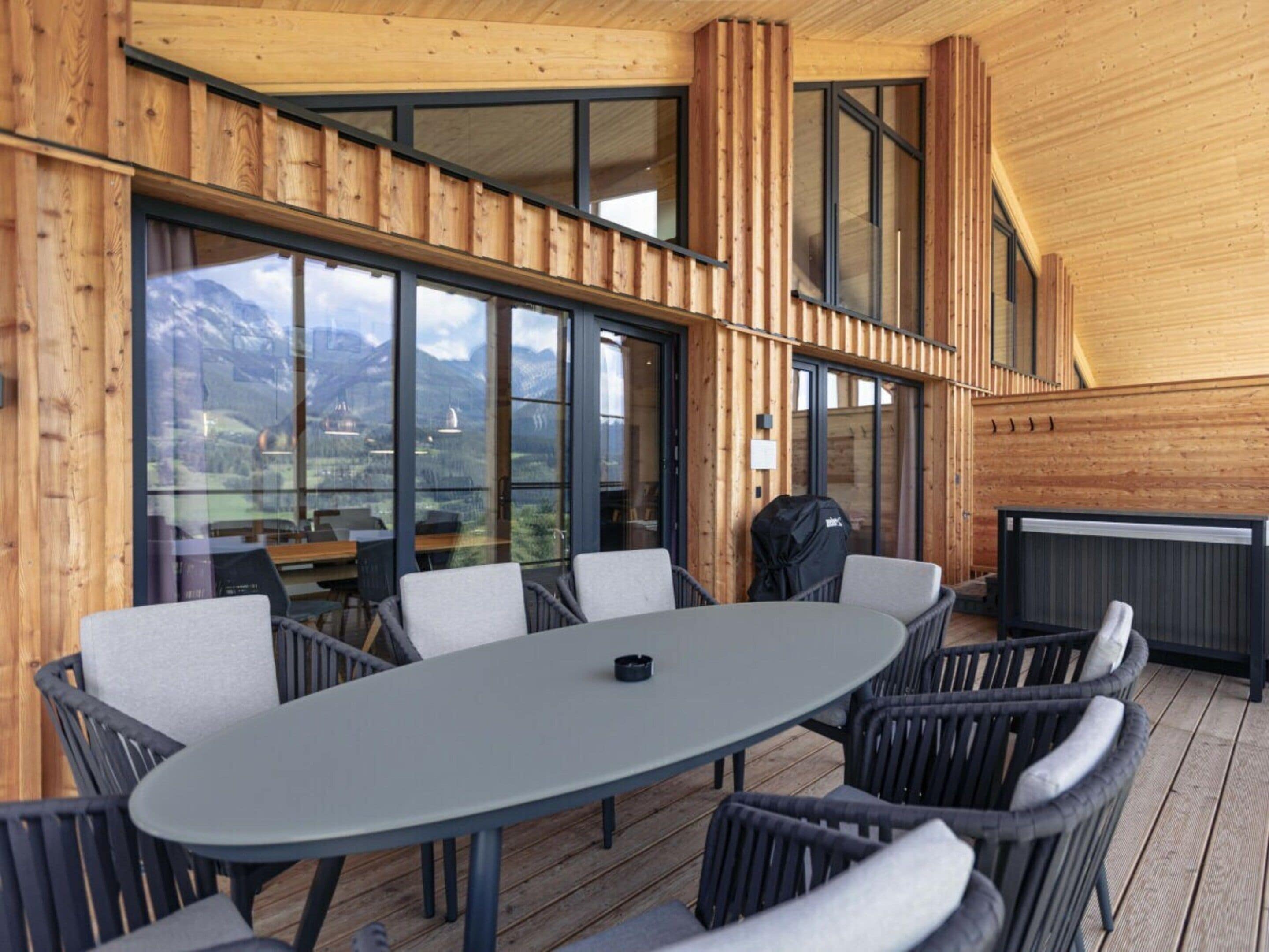 An outdoor dining area with a round table and chairs, large windows offering a view of the mountains, and a grill.