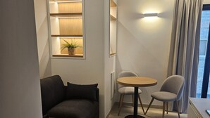 A compact living space with a sofa, a small round table, two chairs, a potted plant, and a window with curtains. - Fahana Residences Arguineguín (Arguineguín)