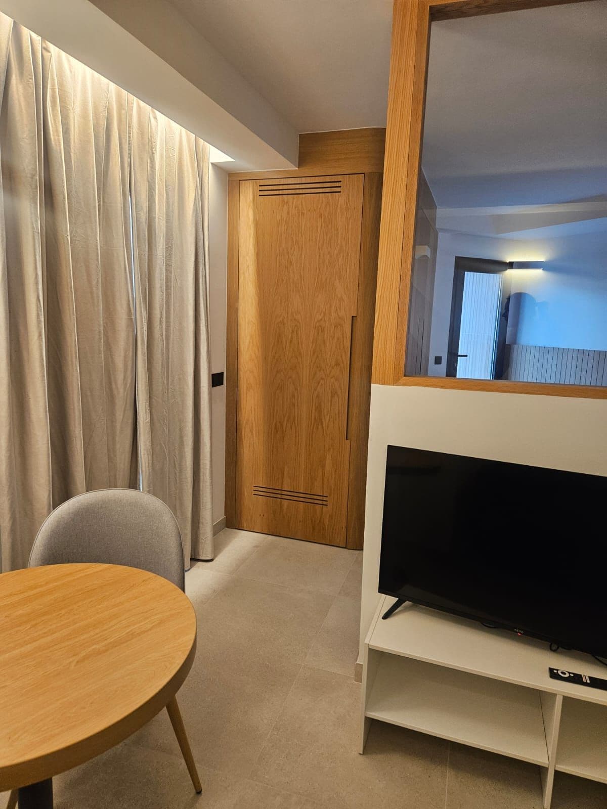 A hotel room with a wooden door, a television on a stand, a round table, and a window with curtains.