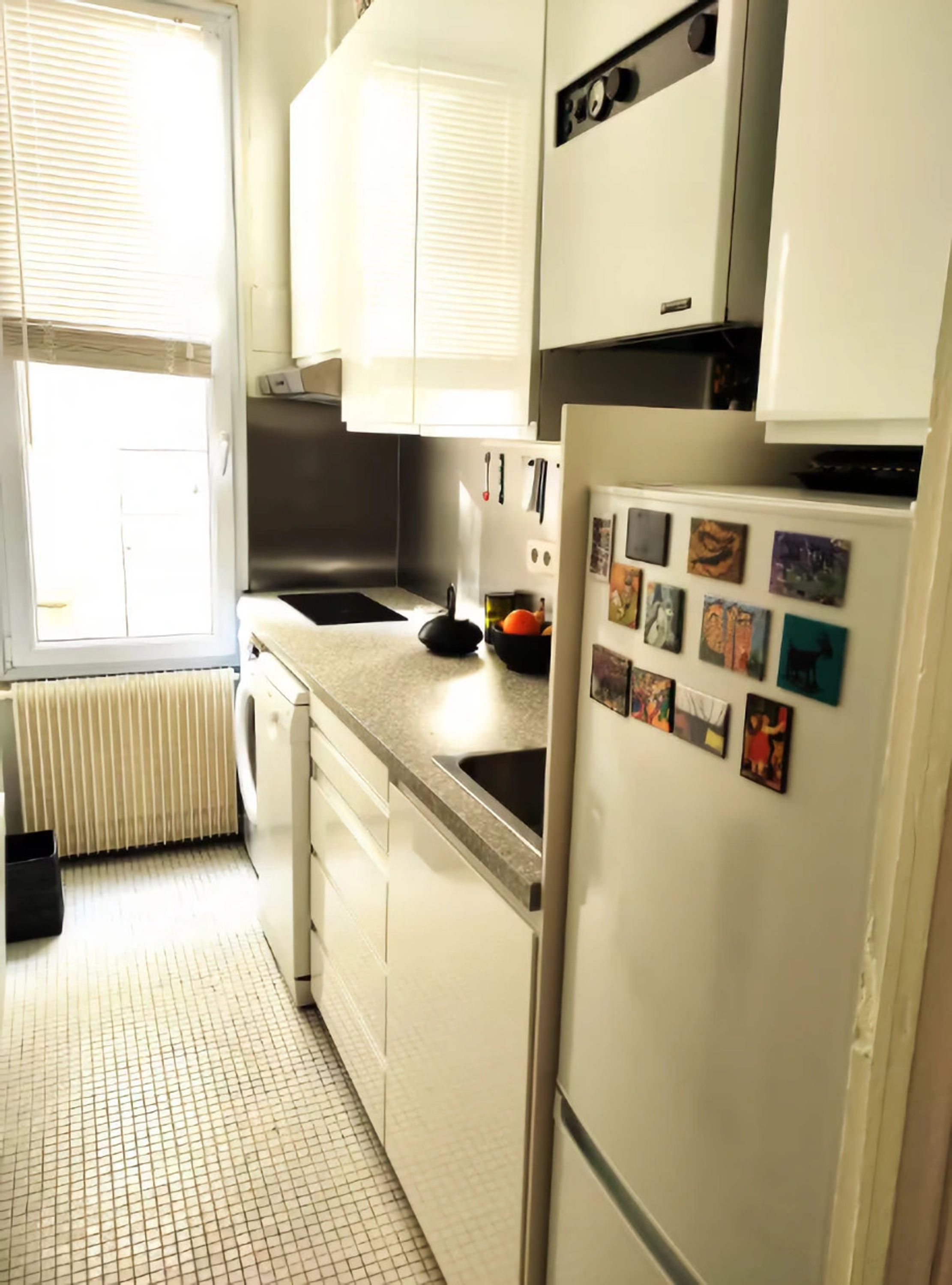 Una cocina con gabinetes blancos, un lavamanos de acero inoxidable, un refrigerador con imanes y una ventana con persianas.