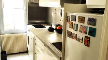 Una cocina con gabinetes blancos, un lavamanos de acero inoxidable, un refrigerador con imanes y una ventana con persianas.