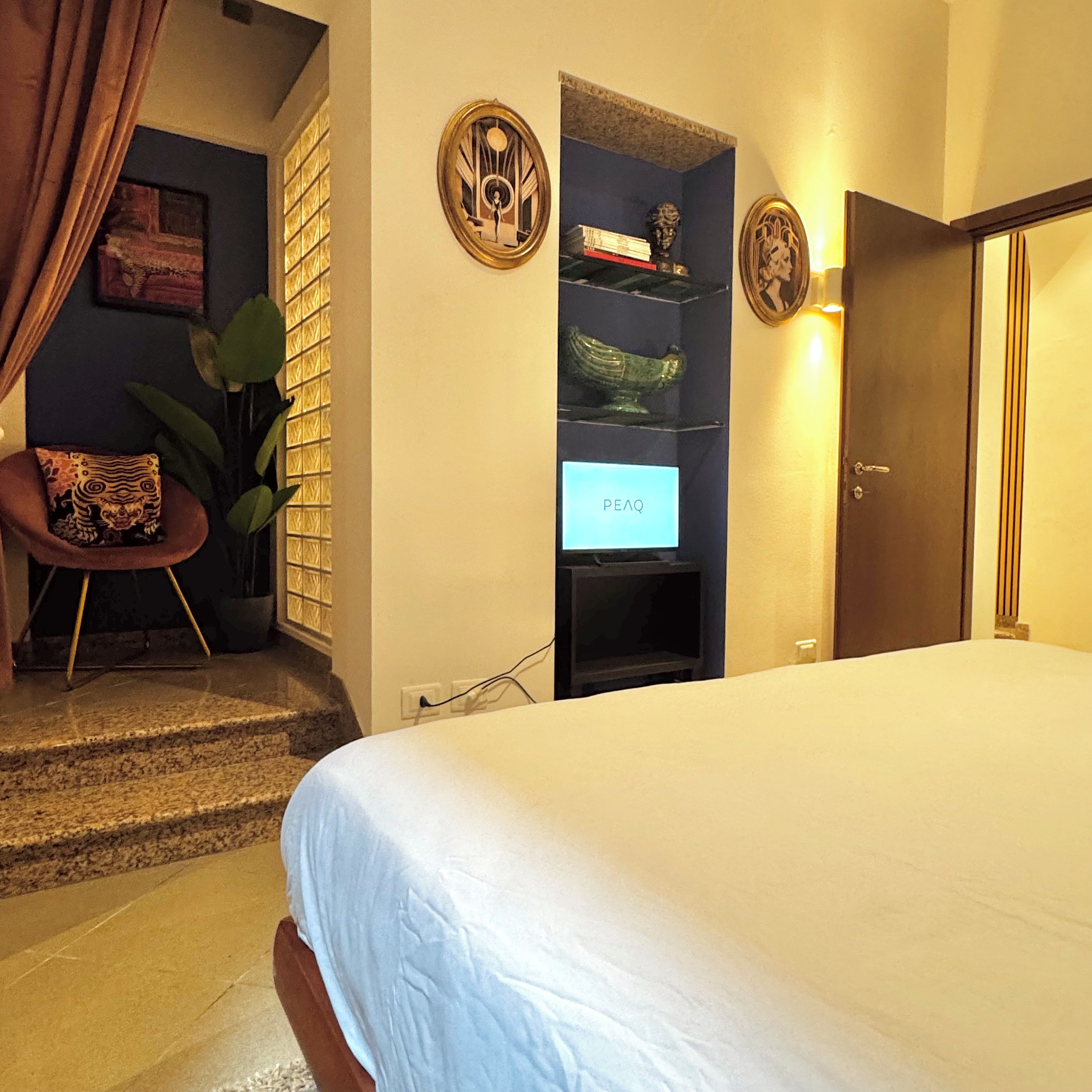 A hotel room with a bed, a seating area with a chair and a plant, a shelf with decorative items, and a door.
