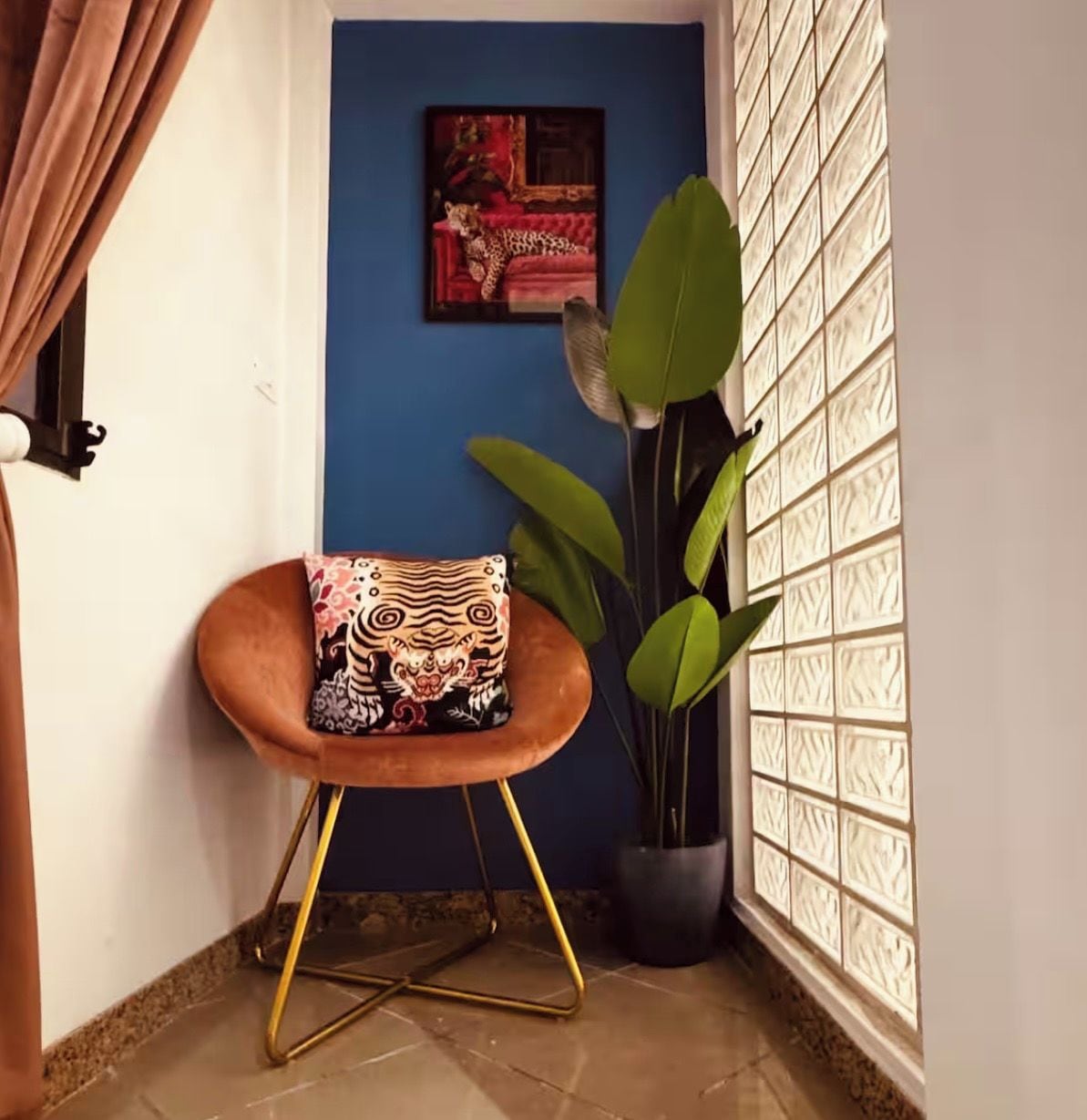 Ein Eckzimmer mit blauer Wand, einem gerahmten Bild, einem Blumentopf, einem gepolsterten Hocker und einem Fenster mit gemusterten Jalousien.