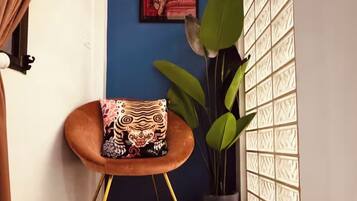 Ein Eckzimmer mit blauer Wand, einem gerahmten Bild, einem Blumentopf, einem gepolsterten Hocker und einem Fenster mit gemusterten Jalousien.