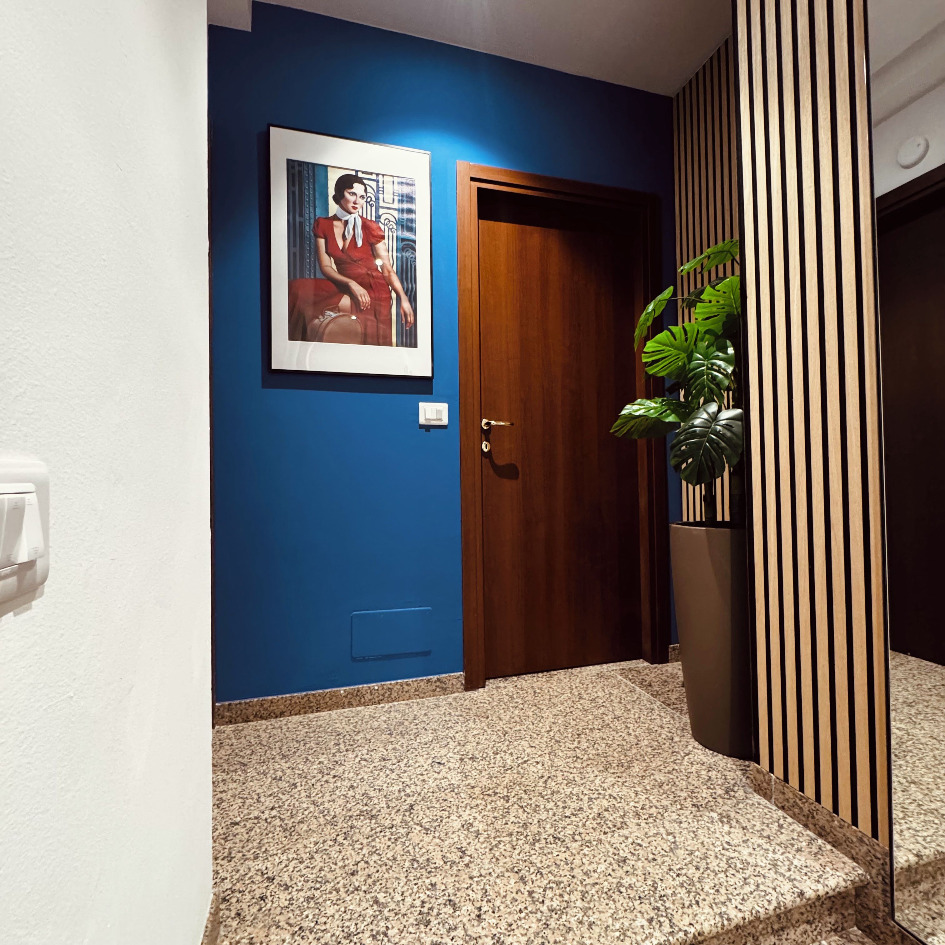 A hallway with a blue wall, a wooden door, a framed picture, a potted plant, and a light switch.
