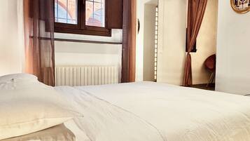 Ein Schlafzimmer mit einem Bett, einem fensterbelaubten Fenster, einem Heizkörper und einer Tür zu einem weiteren Raum.