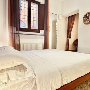Ein Schlafzimmer mit einem Bett, einem fensterbelaubten Fenster, einem Heizkörper und einer Tür zu einem weiteren Raum.