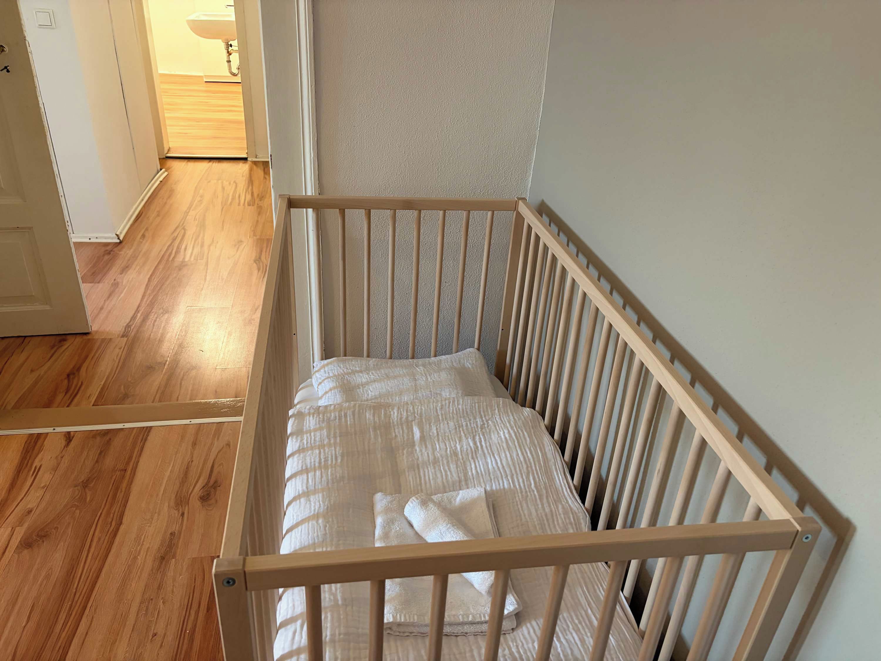 A wooden crib with a mattress and a blanket, positioned against a wall with a wooden floor and a white door in the background.