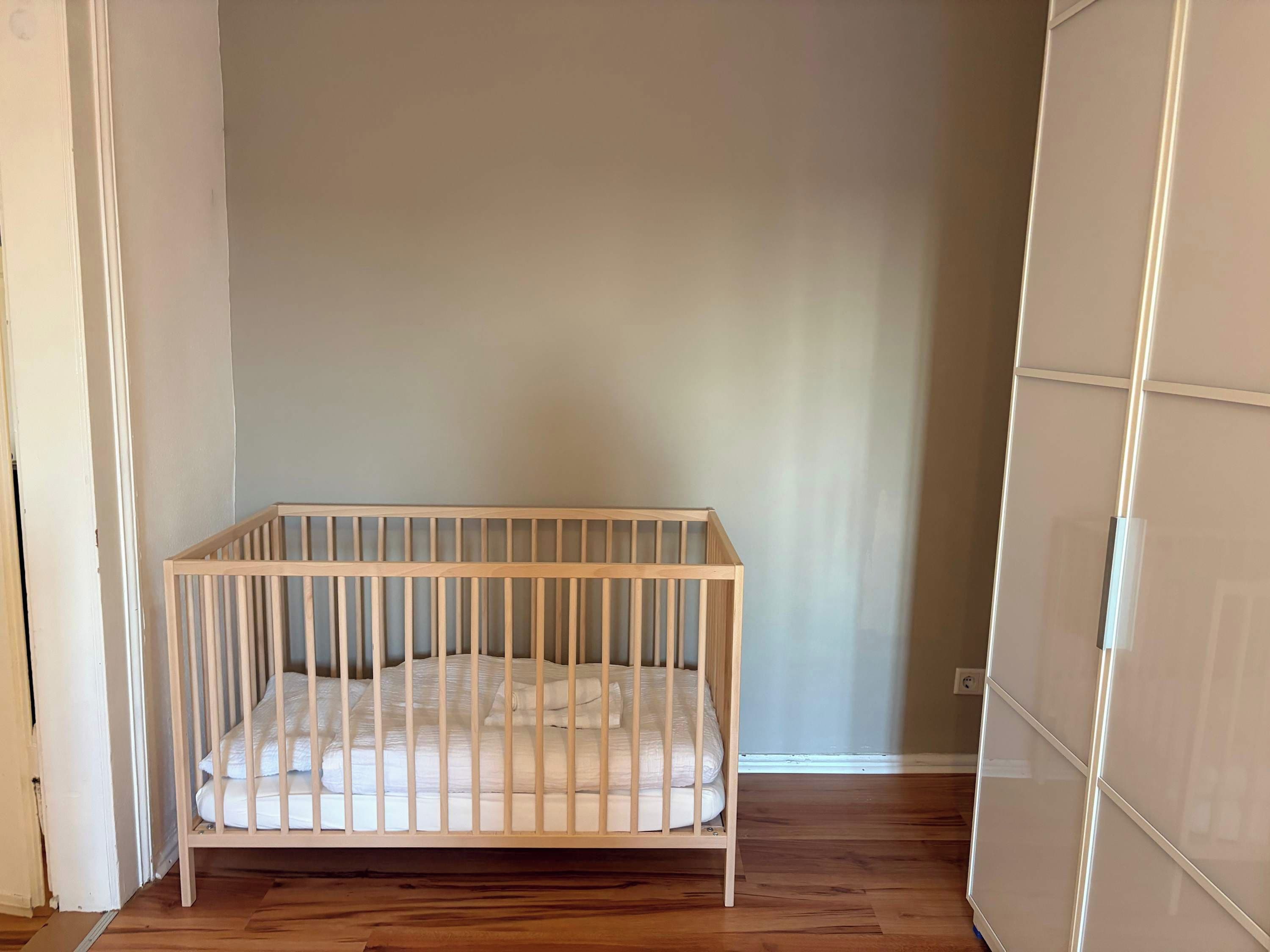 Ein weißes Babybett steht in einer Ecke des Raumes mit Holzboden und einer schlichten Wand.