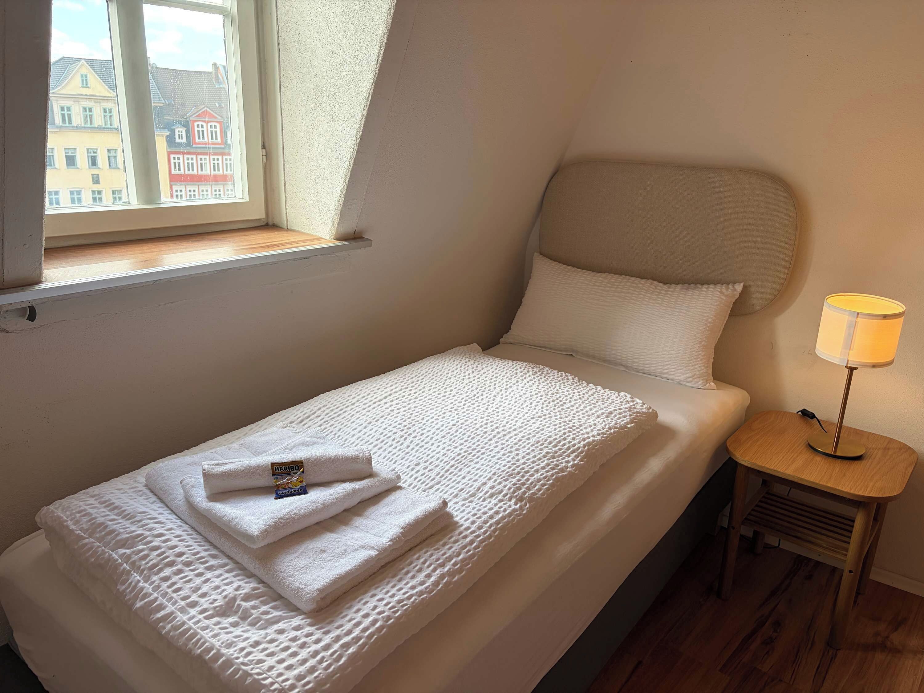 Un letto singolo con una trapuntina bianca e lavorata, un comodino con una lampada e una finestra vista edifici.
