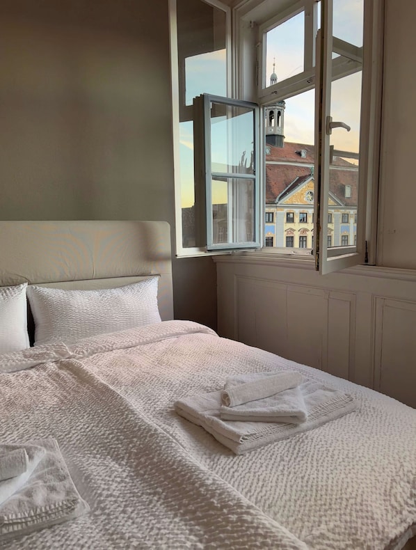 A neatly made bed with a white textured blanket and pillows, a window with a view of buildings, and a white headboard.
