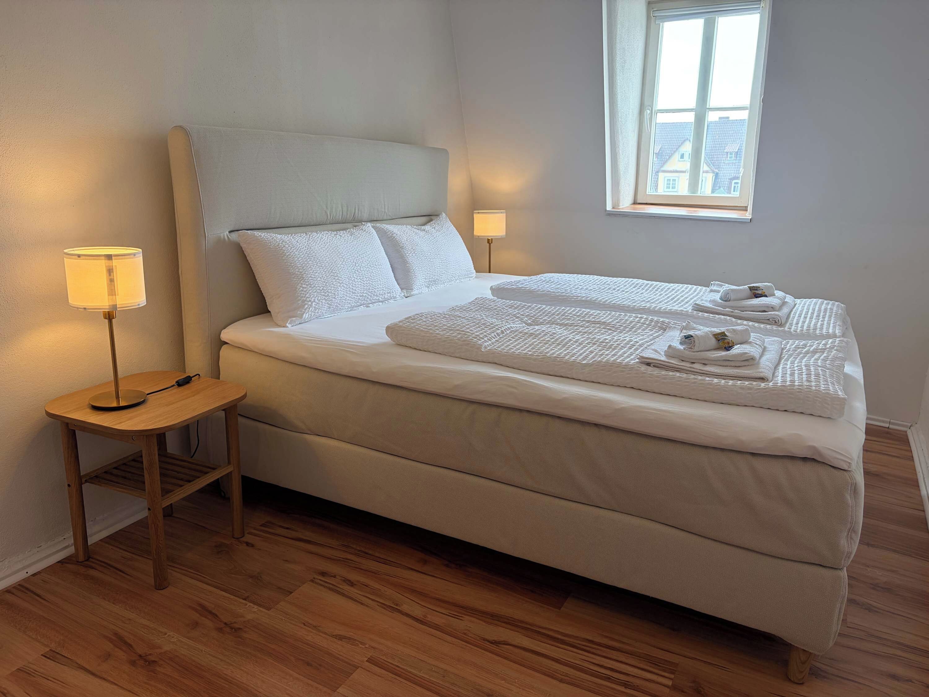 Ein ordentlich bezogenes Bett mit Kopfteil, Nachttischlampe und Handtüchern auf dem Bett.