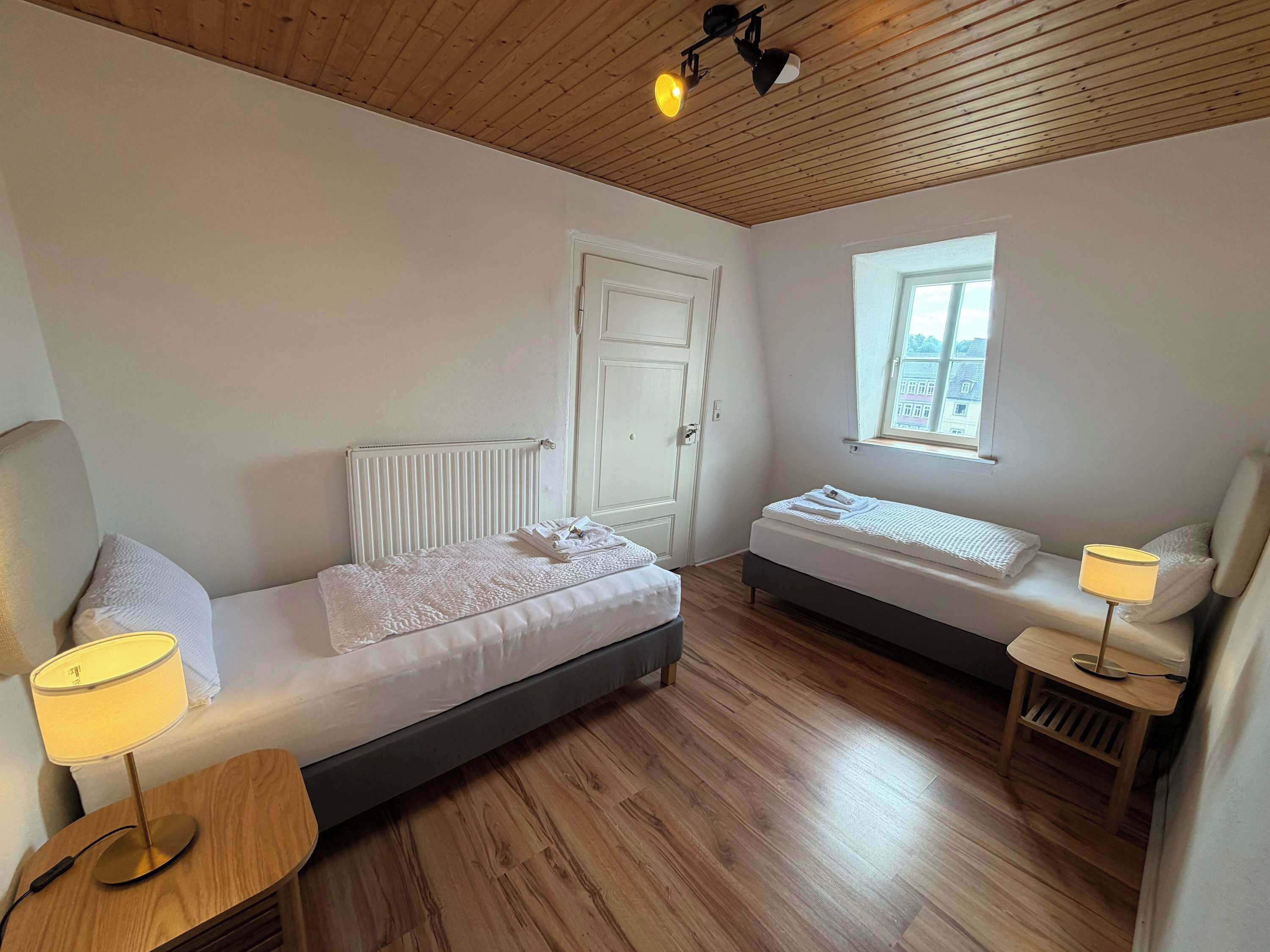 A compact bedroom with two beds, a window, a radiator, and a lamp.