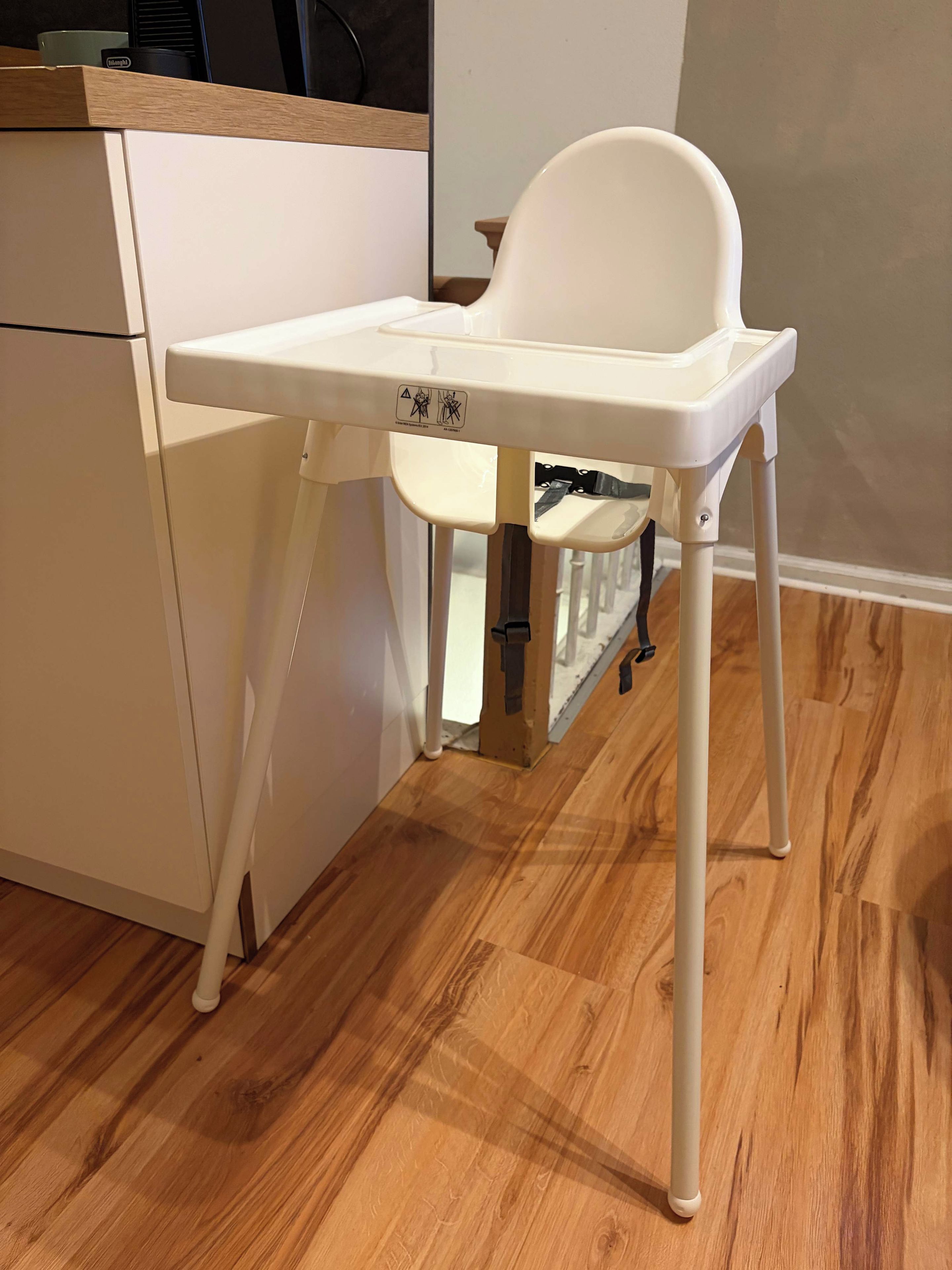 A white high chair with a tray table, positioned in a kitchen area with wooden flooring and a white cabinet.