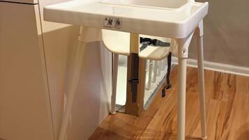 A white high chair with a tray table, positioned in a kitchen area with wooden flooring and a white cabinet.