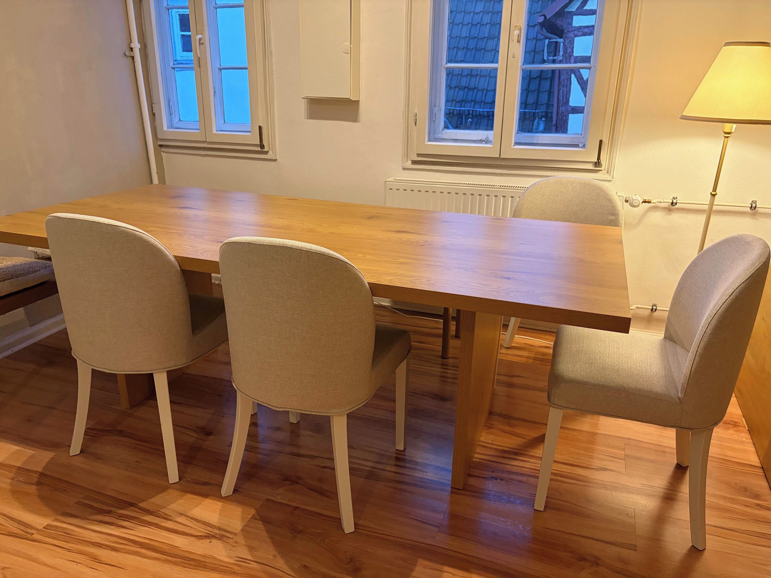 Un coin repas avec une table en bois et quatre chaises, un radiateur et une fenêtre donnant sur un immeuble.