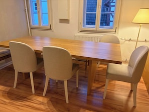 Zona de dinar amb taula de fusta i quatre cadires, un radiador i una finestra amb vista a un edifici.