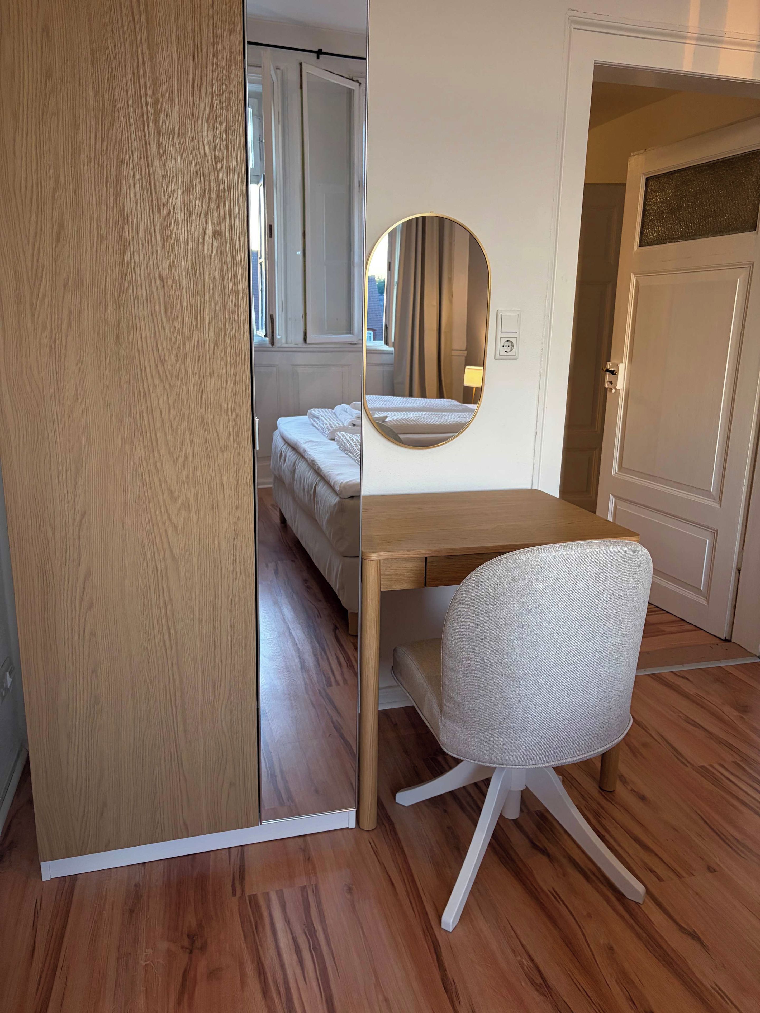 A bedroom with a wooden floor, a bed with white bedding, a round mirror on the wall, and a white chair.