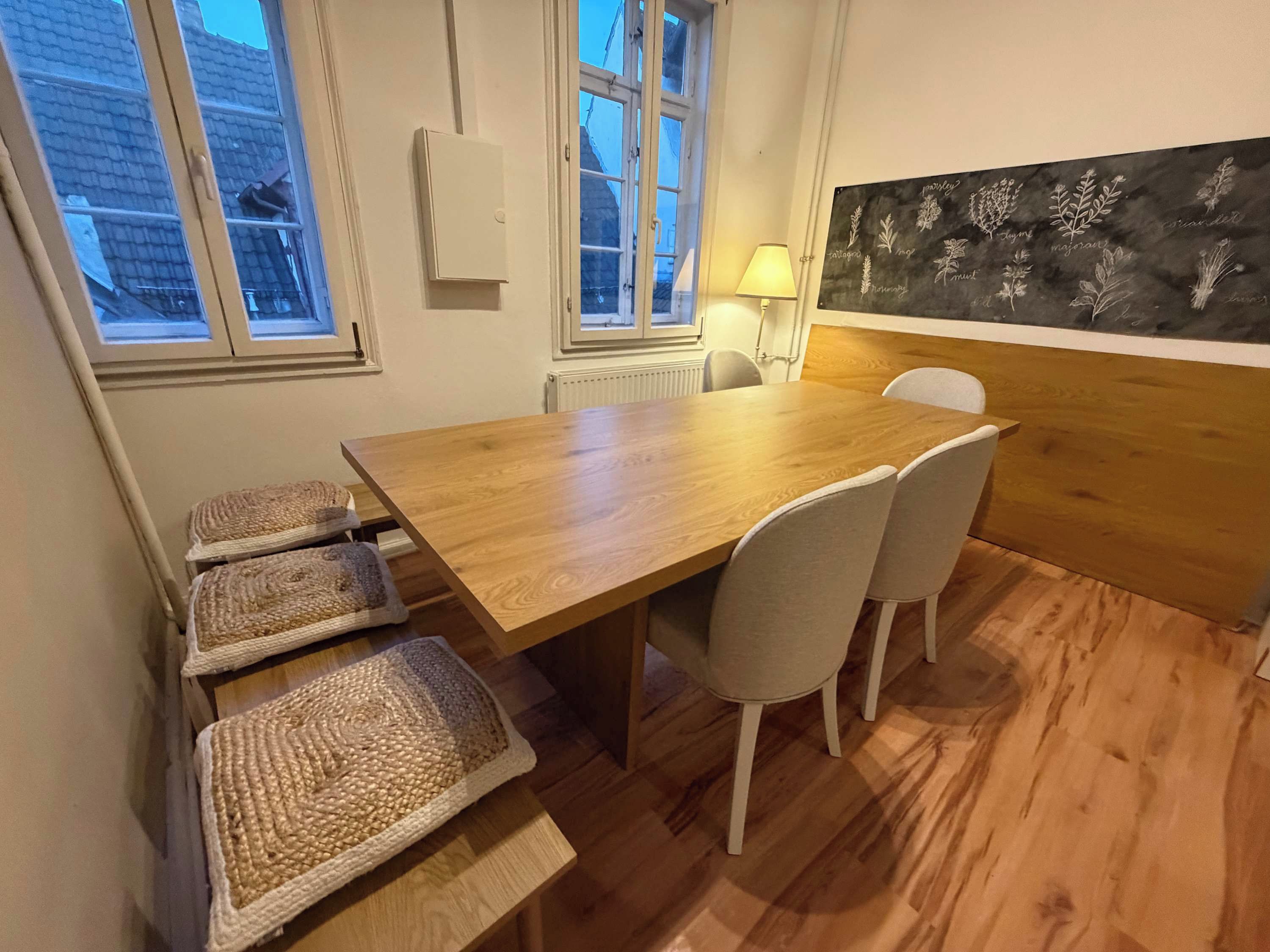 Ein Essbereich mit einem Holztisch, vier Stühlen, einer Wandleuchte und einer Tafel mit Pflanzenabbildungen.